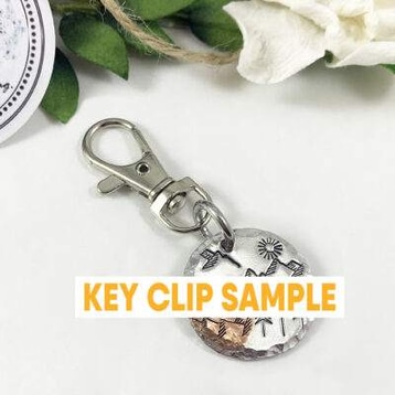 A round metal key clip engraved with mountains, trees, and stars features “KEY CLIP SAMPLE” in bold. Green leaves, a white flower, and a Baby Feet and Heart Necklace are partially visible in the background. Read more at Stamping Cat Studio (stampingcatstudio.com).