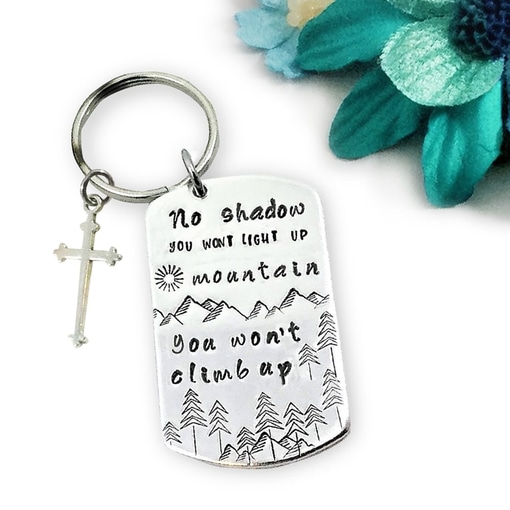 The No Shadow Key 2 is a silver keychain with a cross charm and a tag engraved with mountains, trees, and inspiring words. Blue and teal faux flowers in the background add a vibrant accent. Read more at Stamping Cat Studio (stampingcatstudio.com).
