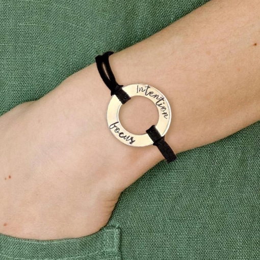 A hand wears a Men Women Personalized Washer Bracelet with a round gold-toned "Intention" and "focus" charm on a black cord. A green textured sleeve appears in the background. Read more at Stamping Cat Studio (stampingcatstudio.com).