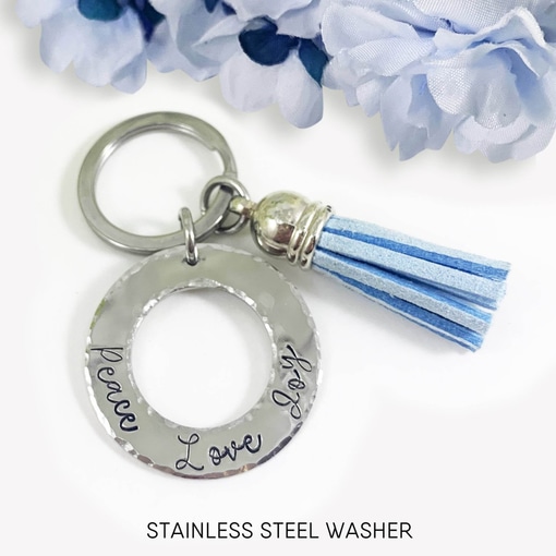 The Personalized Keychain With Color Tassel features a stainless steel washer engraved with "Peace Love Joy" and a blue-and-white suede tassel, set against a background of blue and white artificial flowers. Text below reads: STAINLESS STEEL WASHER. Read more at Stamping Cat Studio (stampingcatstudio.com).