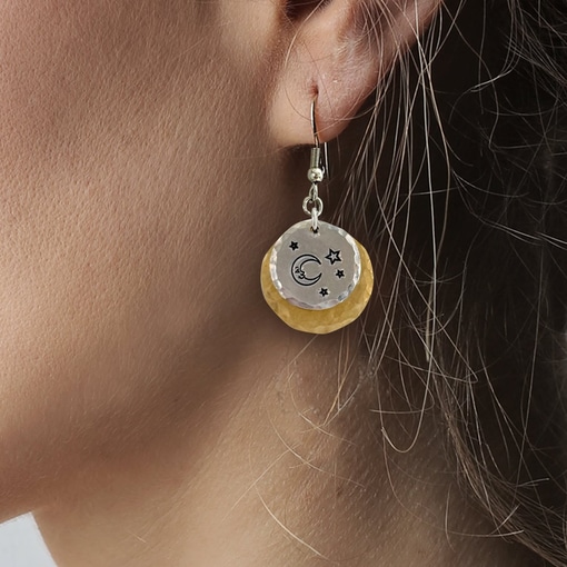 Close-up of a person's ear wearing the Man In The Moon nugold ear—a double-layered dangle earring featuring a silver disc engraved with stars and a crescent moon, layered over a textured gold disc. Brown hair partially frames the ear and earring. Read more at Stamping Cat Studio (stampingcatstudio.com).