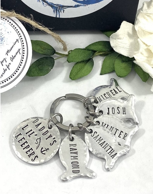 The Daddys Lil Keepers Keyring is silver and features five fish-shaped charms engraved with "RAYMOND," "MICHAEL," "JOSH," and "SAMANTHA." The largest charm reads "DADDY'S LIL' KEEPERS" and includes two small fish, an anchor, leaves, and a white rose. Read more at Stamping Cat Studio (stampingcatstudio.com).