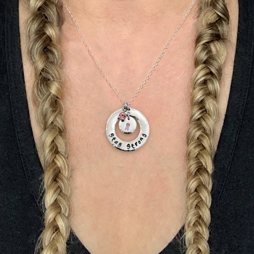 A close-up of a person in a black top with two blonde braids wearing the Stay Strong Pink Ribbon Necklace, featuring a silver pendant engraved with “Stay Strong” and a pink ribbon charm for breast cancer awareness. Read more at Stamping Cat Studio (stampingcatstudio.com).