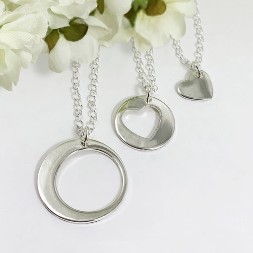 Three necklaces from the Always Family Generations Necklace Set—featuring a large circle, a heart cutout circle, and a small solid heart—hang on silver chains, displayed against white artificial flowers on a white background. Read more at Stamping Cat Studio (stampingcatstudio.com).
