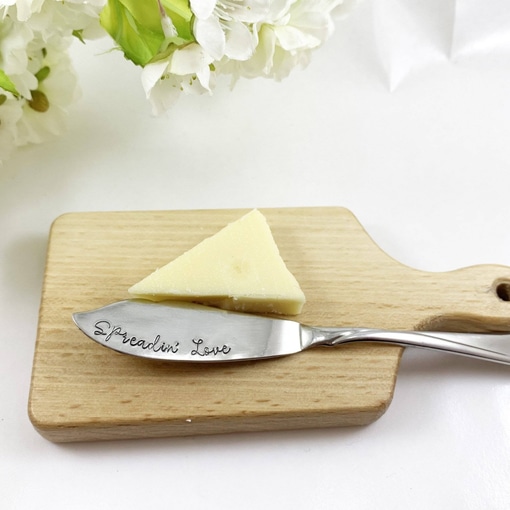 The Spreader Knife and Serving Board Gift Set features a triangular wedge of cheese on a small wooden board, paired with a silver knife engraved “Spreadin’ Love.” White flowers adorn the top left for a fresh, inviting presentation. Read more at Stamping Cat Studio (stampingcatstudio.com).