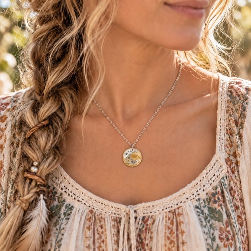 A person with a braided hairstyle wears a Dreamy Sun Moon Stars liifestyle 2 silver necklace, featuring a round pendant with a gold crescent moon and small star cutouts against a floral patterned top. Read more at Stamping Cat Studio (stampingcatstudio.com).