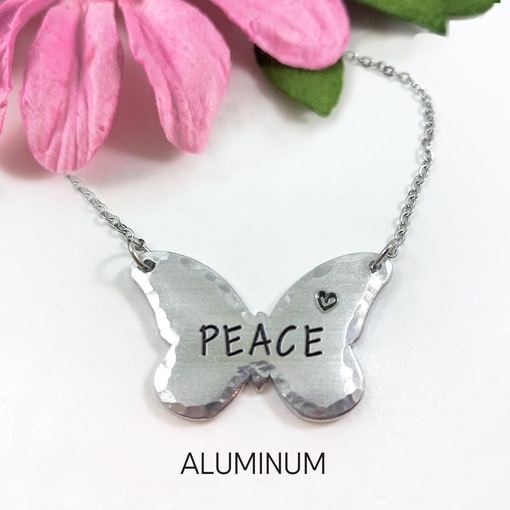 The Inspiring Butterfly Necklace features a silver-toned butterfly pendant engraved with "PEACE" and a heart, hanging from a chain on a white background near a pink flower. The word "ALUMINUM" is displayed below the necklace. Read more at Stamping Cat Studio (stampingcatstudio.com).