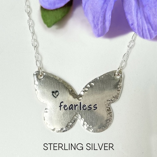 The Inspiring Butterfly Necklace features a hammered sterling silver butterfly on a chain, engraved with a small heart and "fearless" in black. Purple flowers are in the background, with "STERLING SILVER" shown below the necklace. Read more at Stamping Cat Studio (stampingcatstudio.com).