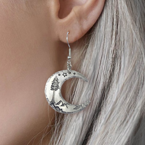 A close-up of a person with long, silver hair wearing Crescent Moon Dreamy Mountain Scene Earrings, featuring engraved stars, mountains, and a pine tree inspired by dreamy landscapes. Read more at Stamping Cat Studio (stampingcatstudio.com).
