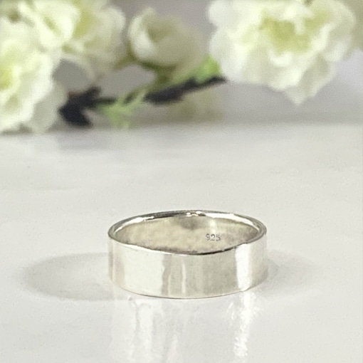 A Personalized Sterling Silver Band, 6mm with a “925” stamp inside rests on a glossy white surface, while soft white flowers and green stems in the blurred background create a delicate, minimalistic atmosphere. Read more at Stamping Cat Studio (stampingcatstudio.com).