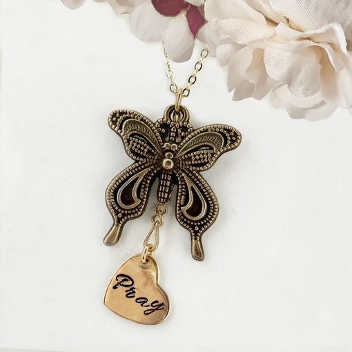 The Bronze Butterfly Neck With Pray Heart Charm main showcases a detailed butterfly pendant alongside a "Pray"-engraved heart charm, displayed against soft white and pale pink artificial flower accents. Read more at Stamping Cat Studio (stampingcatstudio.com).