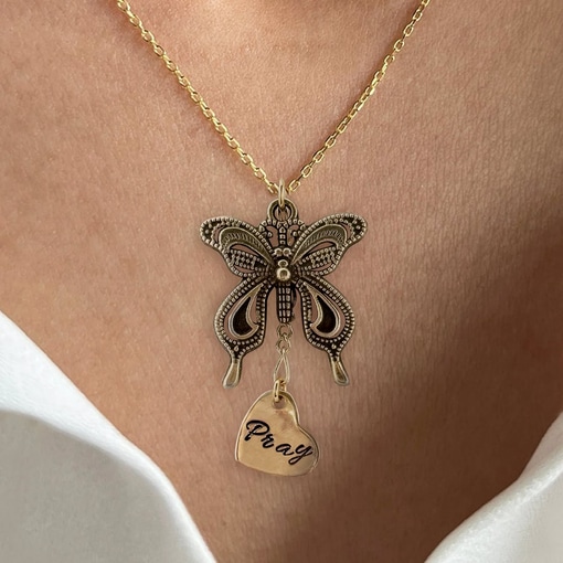 A person in a white off-shoulder top wears the Bronze Butterfly Neck With Pray Heart Charm, featuring a detailed bronze butterfly pendant and a heart charm engraved with "Pray. Read more at Stamping Cat Studio (stampingcatstudio.com).