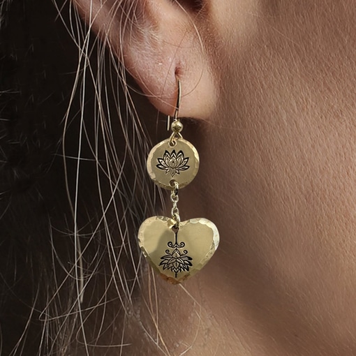 A close-up of a woman's ear wearing Gold Lotus Heart Dangle Earrings, with a lotus-engraved round top and a hammered floral heart pendant linked by a delicate chain. Read more at Stamping Cat Studio (stampingcatstudio.com).