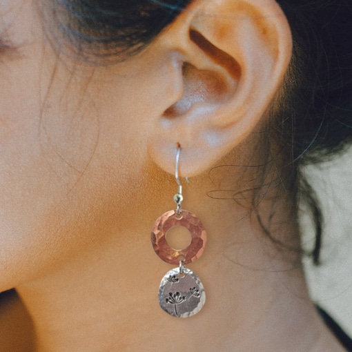 A close-up shot of a woman wearing the Dandelion Dream Dangle ear lifestyle earring: the top features a hammered copper ring, while the bottom is a round silver disk etched with a delicate floral design. Her dark hair is pulled back, and gentle light highlights her skin. Read more at Stamping Cat Studio (stampingcatstudio.com).