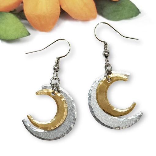 Introducing the Double Crescent Moon Ear Main: a pair of dangling earrings featuring a smaller gold crescent layered over a larger hammered silver crescent, beautifully displayed against a softly blurred background accented with orange and green floral elements. Read more at Stamping Cat Studio (stampingcatstudio.com).