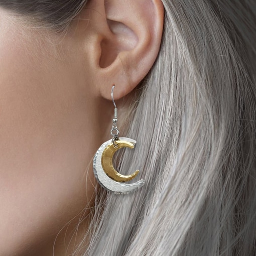 A close-up of a person’s ear adorned with the Double Crescent Moon ear lifestyle earring, featuring long, silvery-gray hair. The dangling earring is crescent-shaped, displaying textured silver on the outside and smooth gold on the inside, while the softly blurred background enhances its elegant design. Read more at Stamping Cat Studio (stampingcatstudio.com).