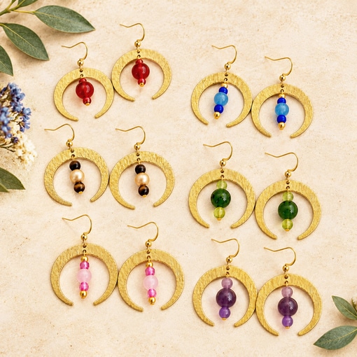 Eight pairs of Boho Crescent Moon Semi-Precious Bead Ear group earrings in crescent shapes with colorful dangling beads are arranged on a textured beige surface with foliage in the corners. Read more at Stamping Cat Studio (stampingcatstudio.com).