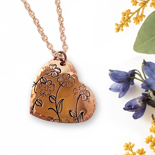 The Heart Bloom Copper Neck main is a hammered copper heart-shaped pendant on a matching link chain, featuring etched line art of three small flowers, displayed on a white background next to sprigs of small blue and yellow flowers. Read more at Stamping Cat Studio (stampingcatstudio.com).