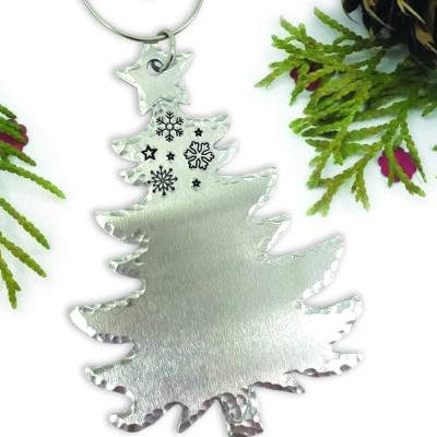 The Christmas Tree Ornament Personalized features engraved black snowflakes and stars near the top, a brushed silver finish, and hammered edges. Faux pine branches and pinecones surround it against a white background. Read more at Stamping Cat Studio (stampingcatstudio.com).