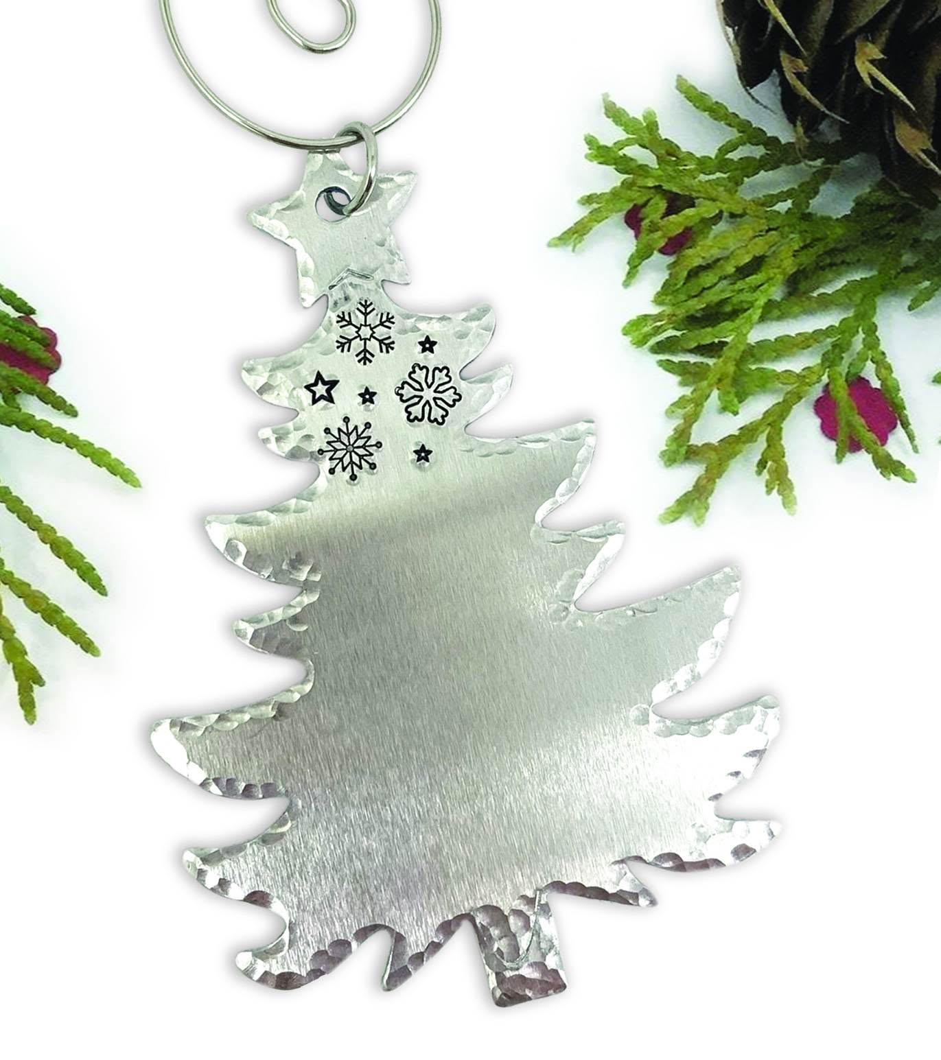 The Christmas Tree Ornament Personalized features engraved black snowflakes and stars near the top, a brushed silver finish, and hammered edges. Faux pine branches and pinecones surround it against a white background. Read more at Stamping Cat Studio (stampingcatstudio.com).