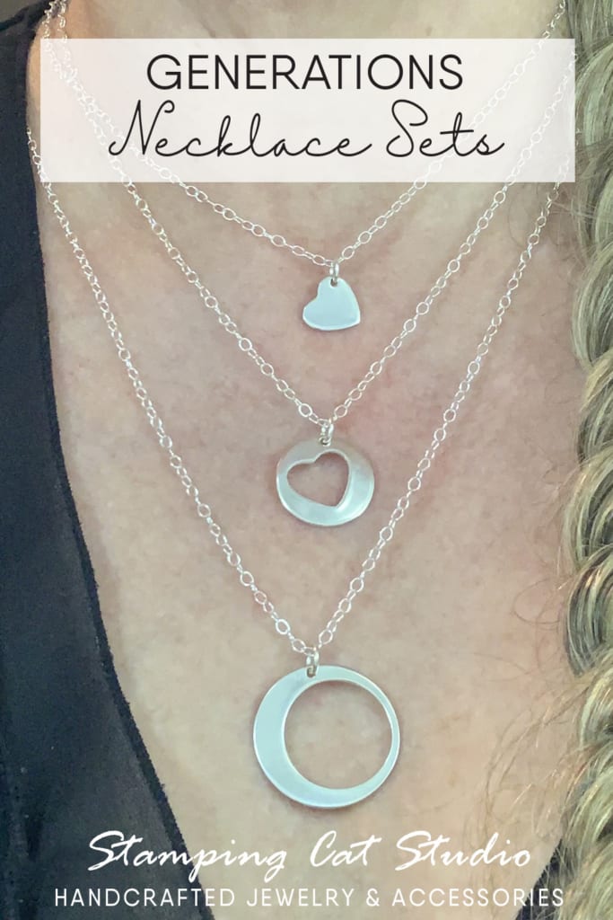 A close-up of a person’s chest wearing three layered silver necklaces: a small solid heart, a medium open heart, and a large open circle—showcasing our handcrafted, handstamped jewelry. "GENERATIONS Necklace Sets" above; “Stamping Cat Studio HANDCRAFTED JEWELRY & ACCESSORIES” below. Read more at Stamping Cat Studio (stampingcatstudio.com).