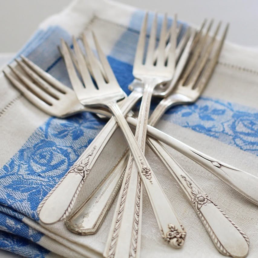 Six vintage silver forks with ornate handles are fanned out on two folded cloth napkins, one with a blue floral pattern, creating a scene that blends classic elegance with subtle boho style for a unique table setting. Read more at Stamping Cat Studio (stampingcatstudio.com).
