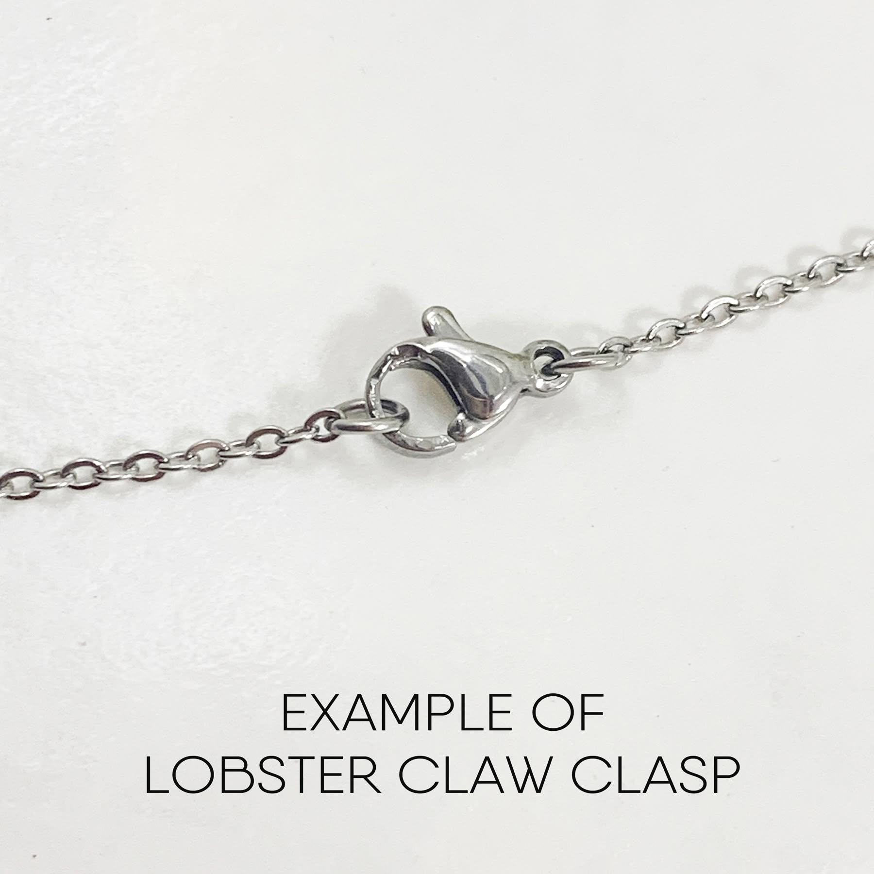 Close-up of the Mystic Moon And Stars Necklace, a silver chain with a lobster claw clasp on a white background. Black text below the chain reads, "EXAMPLE OF LOBSTER CLAW CLASP. Read more at Stamping Cat Studio (stampingcatstudio.com).